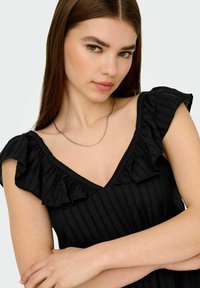 Mujer joven con cabello largo castaño, vistiendo una blusa negra con volantes y un collar de cadena plateada, contra un fondo claro y liso.