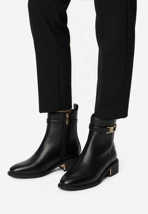 Schwarze Stiefeletten mit seitlichen Reißverschlüssen, dekorativer goldener Schnalle, niedrigen Blockabsätzen, getragen zu schwarzen Hosen.
