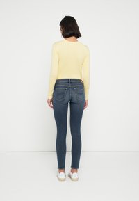 Κολλητό σκούρο μπλε τζιν με πίσω τσέπες, συνδυασμένο με ανοιχτό κίτρινο μπλουζάκι με μακριά μανίκια. Το μοντέλο στέκεται μπροστά σε απλό λευκό φόντο.