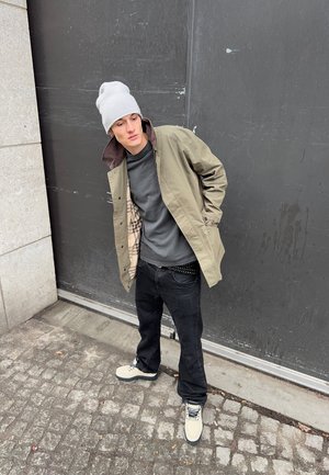 Νέος άνδρας ακουμπά σε έναν σκοτεινό τοίχο, φοράει μπεζ σακάκι, γκρίζο σκουφάκι, μαύρο παντελόνι, γκρι πουκάμισο και λευκά αθλητικά παπούτσια σε έναν λιθόστρωτο δρόμο.