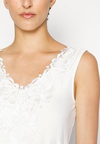 Vestido branco com bordados tonais intrincados ao longo de um decote em V profundo, com textura de tecido suave e design sem mangas.