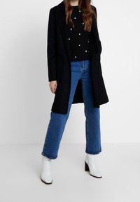 Černý dlouhý kabát přes černý svetr s bílými puntíky, doplněný modrými džínami s širokými nohavicemi a bílými botkami na podpatku. Hladké textury a čisté linie.