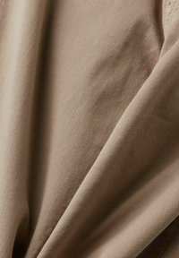 Beige kankaan rakenne, jossa on hienovarainen viiru, esittelee pehmeitä laskoksia ja lempeää laskostumista. Materiaali näyttää kevyeltä ja sileältä.