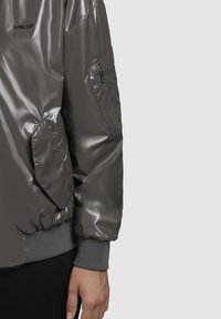 Grijze, glanzende bomberjack met ribgebreide manchetten, ritszak op de mouw en een cirkelvormig detail op het lichaam. Gladde textuur, modern ontwerp.