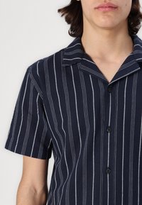 Jeune personne portant une chemise bleu marine à manches courtes avec des rayures verticales blanches et un col à boutons, recadrée au niveau du cou et du bras.