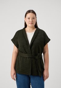 Alyvuogių žalumos siuvinėta striukė su šalikiniu apykakle, trumpomis rankovėmis ir juosmeniu raiščiu, dėvima ant baltos bluzės, derinama su mėlynomis džinsomis.