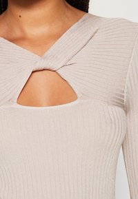 Gros plan sur un haut en maille beige clair côtelé avec un tissu torsadé et une découpe triangulaire au niveau de la partie supérieure de la poitrine, porté par une personne.