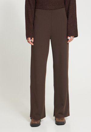 Personne portant un pantalon droit marron foncé, un pull marron, des bagues aux doigts et des chaussures marron sur un fond clair uni.