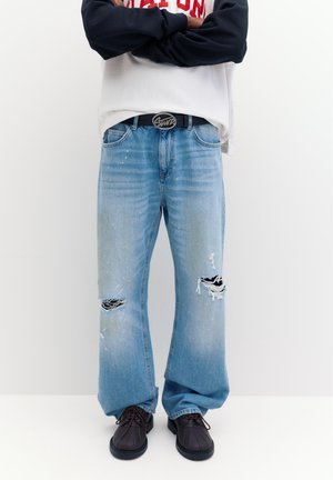 Personne portant un jean bleu clair déchiré à jambes larges, une ceinture noire avec une boucle "SWC", des chaussures noires, et une chemise à manches longues blanche et noire.