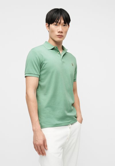 Νέος άνδρας στέκεται με το πρόσωπο προς τα εμπρός φορώντας πράσινο κοντομάνικο πόλο και λευκό παντελόνι με το ένα χέρι στην τσέπη, μπροστά σε απλό φόντο.