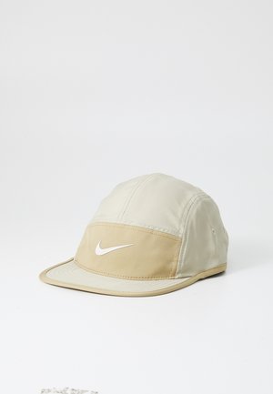 Beige polyester kasket med en beige frontpanel, et hvidt Nike-logo og en buet skygge. Har syninger og ventilationshuller.
