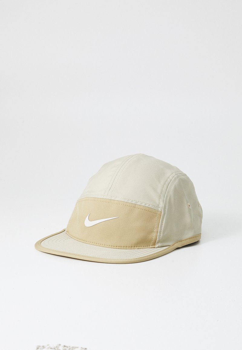Beżowa czapka poliestrowa z beżowym przednim panelem, białym logo Nike oraz zakrzywionym daszkiem. Zawiera detale w postaci przeszyć oraz oczka wentylacyjne.