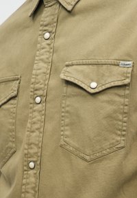 Olívazöld pamut ing, hegyes gallérral, elöl patentgombokkal és két mellzsebbel, amelyek fedéllel rendelkeznek.