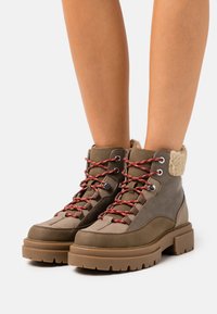 Stivali da trekking marroni con suola spessa, realizzati in materiale testurizzato, con lacci rossi e gialli a contrasto e un colletto morbido di colore crema.