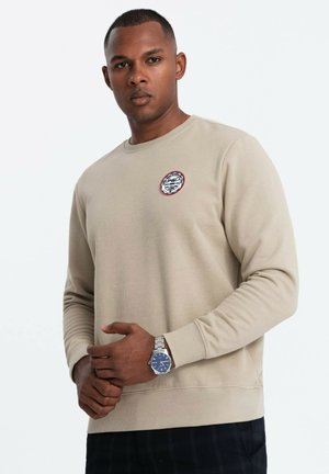 Beige sweatshirt lavet af struktureret stof, med et rundt patch-design på brystet og ribbede ribkanter, parret med et ur.