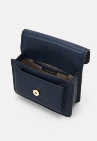 Námorně modrá kožená křížová taška s texturovaným povrchem, zlatým zacvakávacím uzávěrem, otevřenou přihrádkou a štítkem uvnitř.