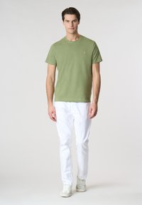 T-shirt verde oliva a maniche corte, colletto rotondo, abbinato a pantaloni bianchi e sneakers chiare. Look semplice e casual con una minimal branding.