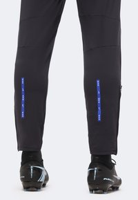 Κάτω άκρα με μαύρο παντελόνι Nike με μπλε κάθετα λογότυπα Nike και μαύρα παπούτσια ποδοσφαίρου Nike Air Zoom Mercurial σε λευκό φόντο.