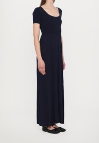 Marineblaues Maxikleid mit kurzen Ärmeln, geripptem Oberteil und fließendem, plissiertem Rock. Kombiniert mit schwarzen flachen Schuhen mit dekorativen Akzenten.