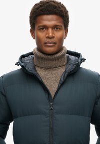 Donker teal pufferjack met een rits aan de voorkant, gecombineerd met een lichtbruine ribgebreide coltrui. Gladde stoftextuur met een aansluitend ontwerp.