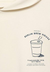 Hanorac crem cu un grafic albastru reprezentând o cană de cafea cu aburi, etichetat "Berlin Brew House" și detalii despre locație dedesubt.