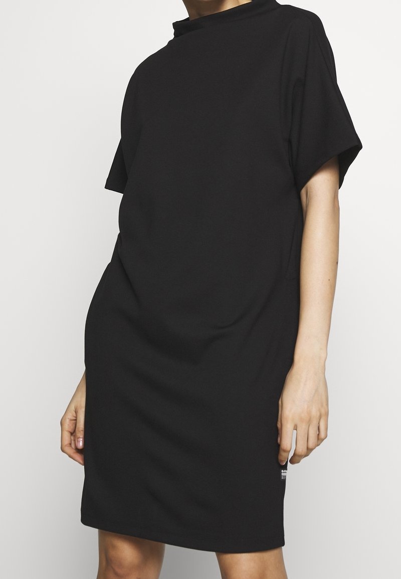 Femme portant une robe noire ample à manches courtes, debout sur un fond blanc uni.