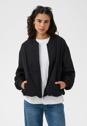 Femme portant une veste noire zippée par-dessus un t-shirt blanc et un jean bleu, avec des lunettes de soleil posées sur sa tête et les mains dans les poches de la veste.