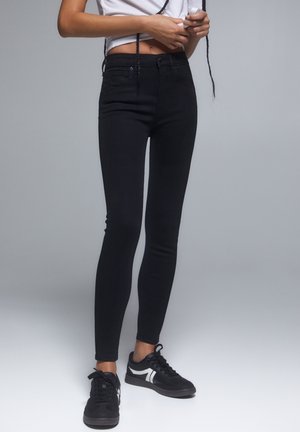 Personne portant un jean skinny noir, des baskets noires et blanches, et un top blanc court, debout devant un fond gris uni.