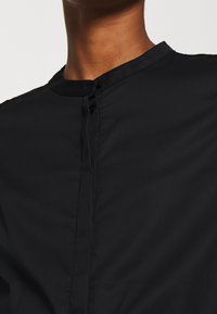 Camicia nera con colletto alto, chiusura a bottoni e texture liscia, senza motivi o accentuazioni visibili.