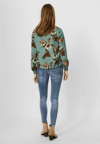 Bluse med blomstermønster i teal med elastiske manchetter ved ærmerne, kombineret med tætsiddende blå jeans og beige ankelstøvler set fra en models ryg.