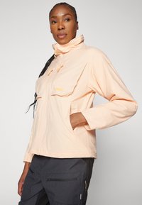 Veste imperméable pêche clair avec un col haut, deux grandes poches avant et fermeture zip. Texture lisse avec des détails de cordon ajustable.