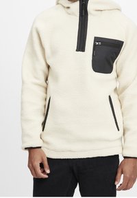 Fleece pullover hoodie in crème met een zwarte borstzak en zijzakken, voorzien van een kwart-ritssluiting en een textuurstof.