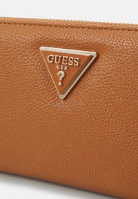 Portafoglio marrone con texture caratterizzato da un logo triangolare in metallo di Guess, con il nome del brand e un dettaglio a forma di punto interrogativo. Chiusura con zip nella parte superiore.