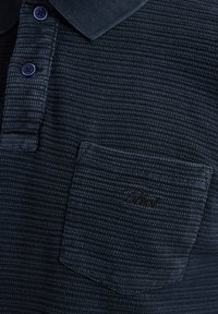 Polo cu culoare bleumarin, realizat dintr-un material texturat, cu deschidere la gât cu trei nasturi și un buzunar pe piept cu un logo brodat. Guler și manșete cu nervuri.