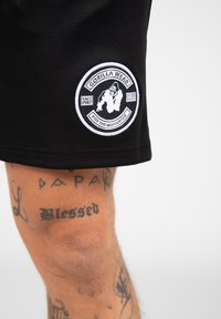Zwarte shorts van rekbare stof hebben een ronde witte patch met het "Gorilla Wear"-logo en tekst, gestikt nabij de zoom.