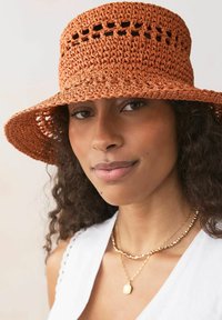 Sombrero de paja de ganchillo en color óxido con un diseño de tejido abierto y un ala ancha. Combinado con cadenas de oro en capas y una blusa blanca.