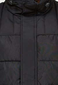 Svart vadderad jacka med hög krage, sömmar i paneler och en vertikal framfick med knappdetaljer. Slät, matt yta.