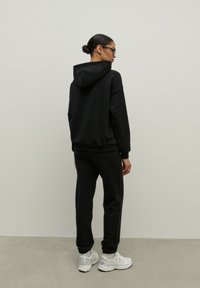 Čierna mikina s kapucňou a sťahovacou šnúrkou, v kombinácii s čiernymi teplákmi so stranovými pruhmi. Oblečené s bielymi športovými topánkami.