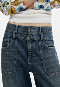 Jean en denim bleu foncé, avec une fermeture à double bouton, de grandes poches avant et une texture légèrement décolorée. Conçu avec une taille mi-haute.