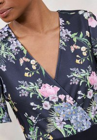 Cvjetna majica s omotom u tamnoplavoj boji, s višebojnim cvjetnim uzorcima, kratkim rukavima i dubokim V-izrezom. Glatka satenasta tekstura.