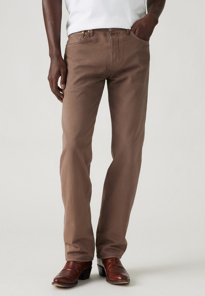Homme portant un pantalon marron coupe droite avec des chaussures en cuir marron et une chemise blanche, debout avec une main dans la poche sur un fond uni.