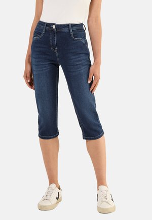 Jean capri en denim bleu foncé avec une taille ajustée, des poches avant et des ourlets retroussés. Assortis à des baskets blanches avec des accents noirs.