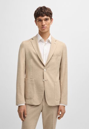 Light beige blazer made of textured fabric, featuring two front pockets and two buttons. Worn over a white shirt, has a tailored fit.