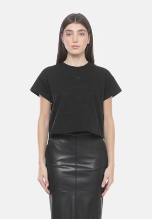 Femme aux longs cheveux bruns portant un t-shirt graphique noir à manches courtes et une jupe crayon en cuir noir, debout devant un arrière-plan uni.