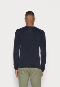 Maglione a maniche lunghe di colore blu navy in maglia con un motivo texturizzato, scollo rotondo e maniche raglan, abbinato a pantaloni verdi.