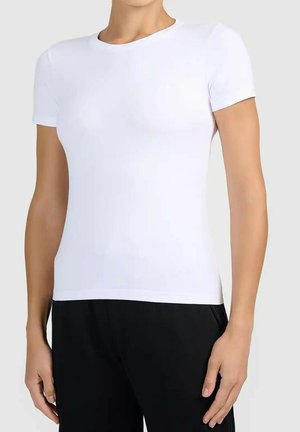 Osoba ubrana w dopasowany biały t-shirt z krótkim rękawem i okrągłym dekoltem oraz czarne spodnie, stojąca na jednolitym jasnym tle.