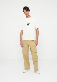 White graphic t-shirt with coffee pot design, paired with beige cargo pants featuring side pockets and a relaxed fit. White sneakers.