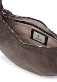 Grå mocka-väskas insida med silverdragkedja, med ett beige mönstrat foder och en etikett som visar varumärket. Enkel, minimalistisk design.