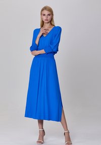 Robe longue bleue à manches longues avec un décolleté en V, accents froncés aux poignets et taille smockée. Comprend une fente sur le côté et une silhouette fluide.