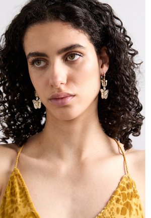 Vrouw met krullend donker haar draagt gouden vlinderoorbellen en een mosterdgele fluwelen jurk met dunne bandjes, kijkt iets naar links.